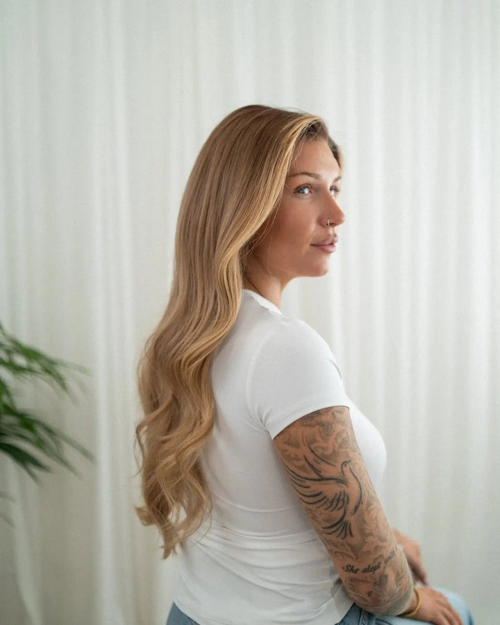 Woman with tattoos wearing a white shirt and blue jeans against a light curtain background