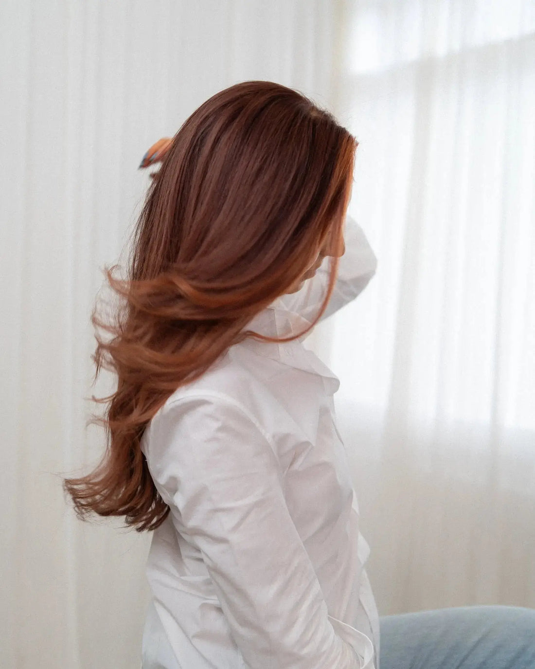 Person with long brown hair wearing a white shirt and blue jeans against a white curtain background