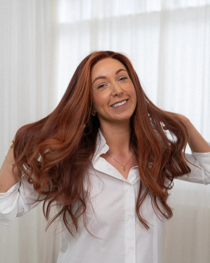 Woman with long red hair wearing a white shirt against a white curtain background