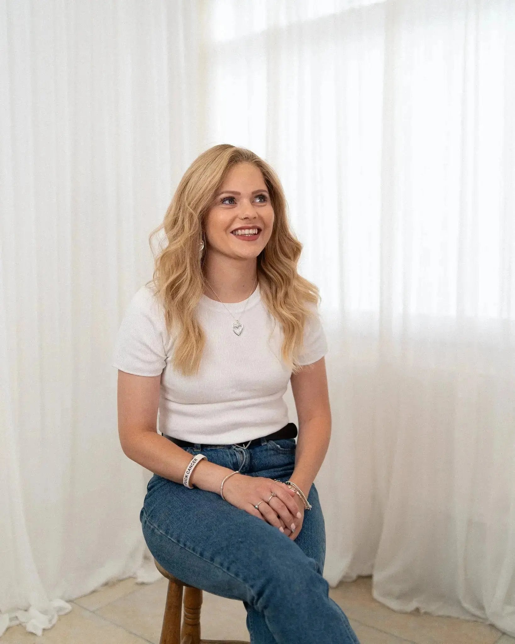Woman sitting on a stool wearing a white t-shirt and blue jeans against a white curtain background