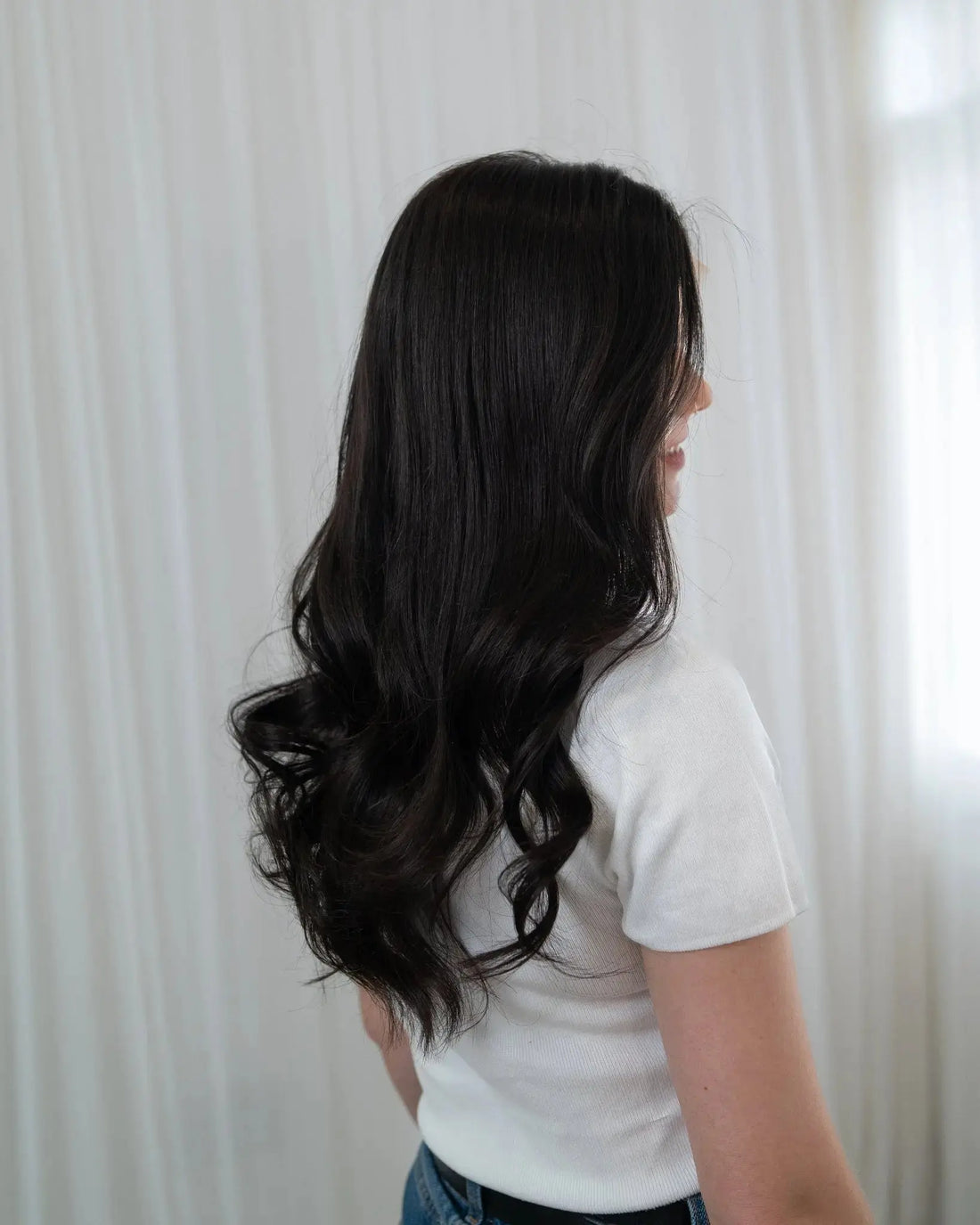 Person with long, wavy black hair wearing a white shirt and blue jeans against a white curtain background